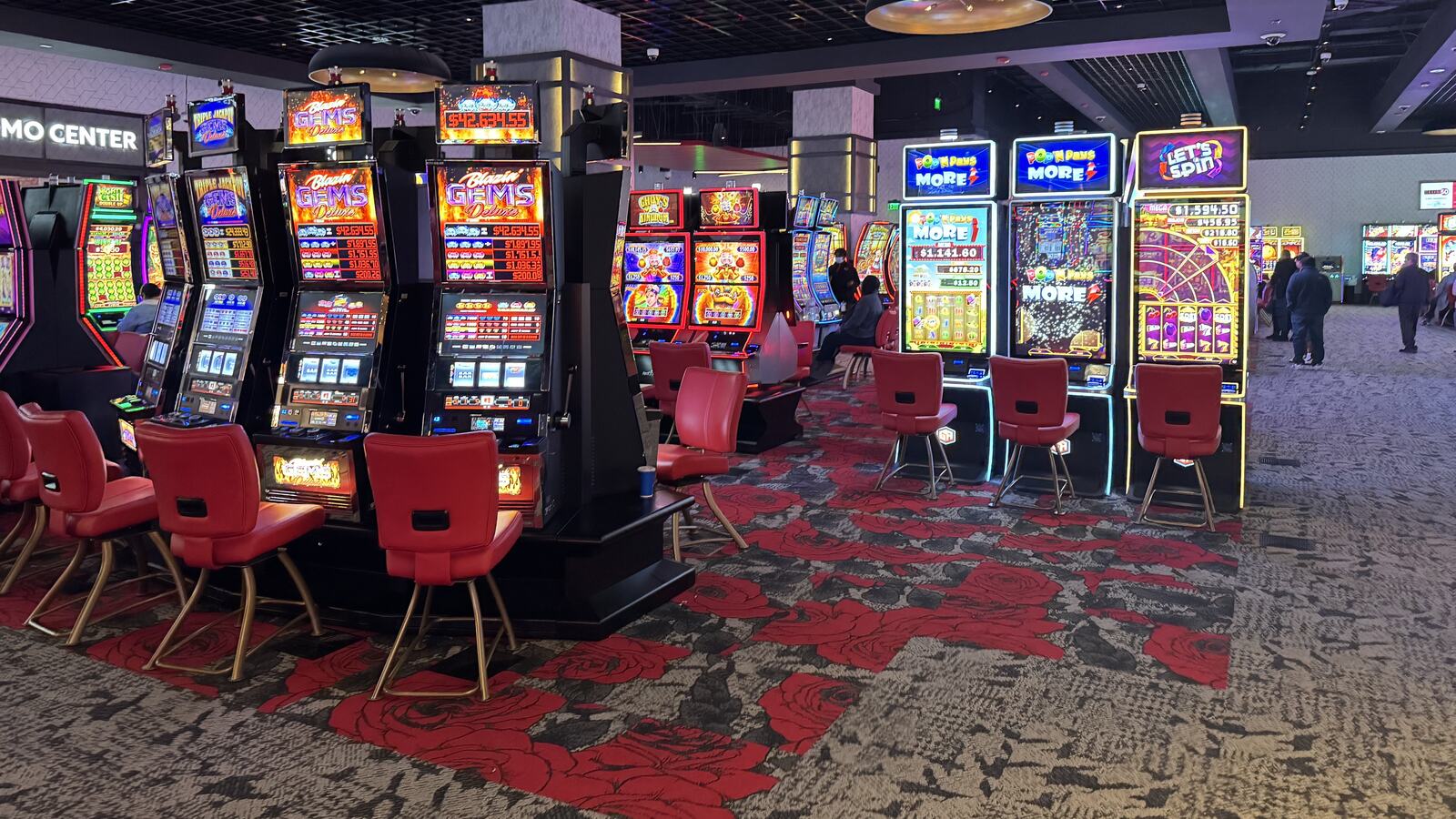 Rad av upplysta spelautomater med färgglada skärmar på ett casinogolv, med röda stolar framför varje maskin